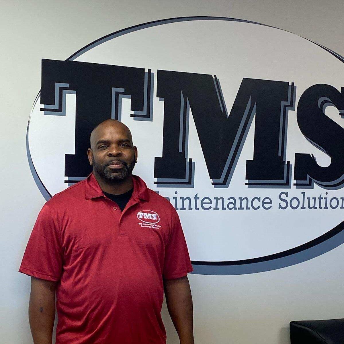a man smiling and standing in front of the TMS logo on the wall behind him